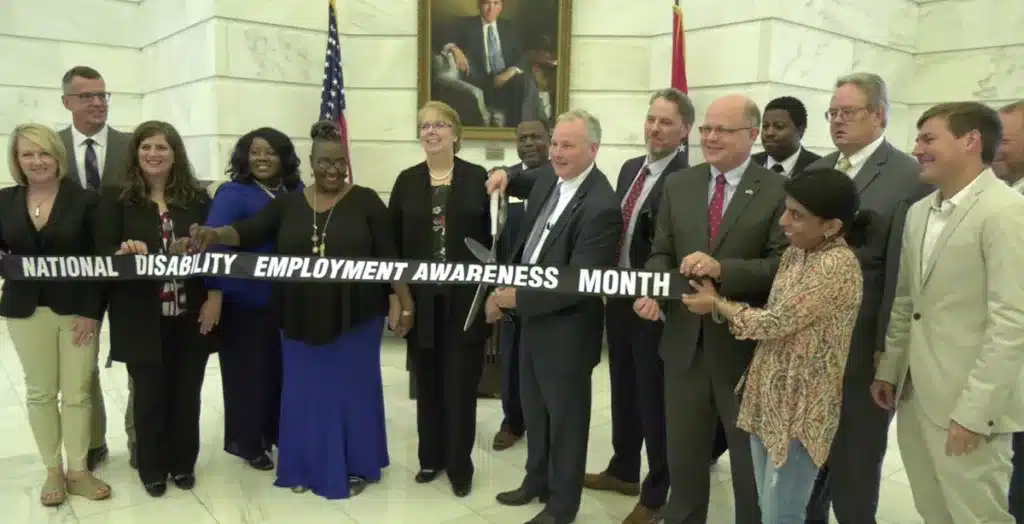 [Photo Description: This is an image of a ribbon cutting ceremony in the Arkansas State Capitol building. A group of women and men are standing in a line, holding a black ribbon that says "National Disability Employment Awareness Month" in white letters, while a man in the middle of the group holds a large pair of scissors near the ribbon.]